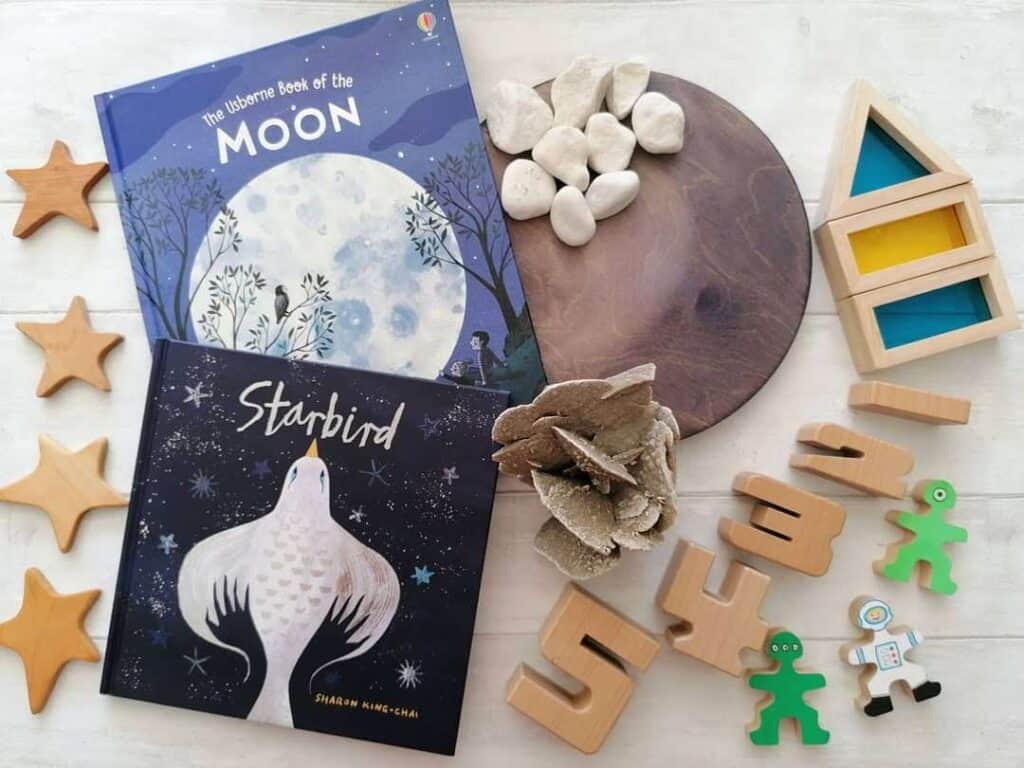 Starbird book flatlay with the Usborne Book of the Moon, four wooden stars to the left of the book. A hand stained moon wooden disc with white rocks and desert rose rock, to the right are the numbers 1 to 5 using SumBlox blocks. Flockmen (small wooden people) painted as two aliens and an astronaut with blue and yellow rainbow blocks to form a rocket.