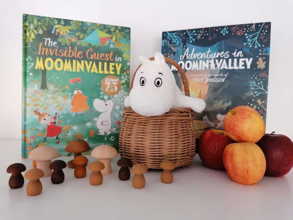 The Invisible Guest in Moominvalley book with character, apples and wooden muchsrooms. Moomintroll sits in a basket