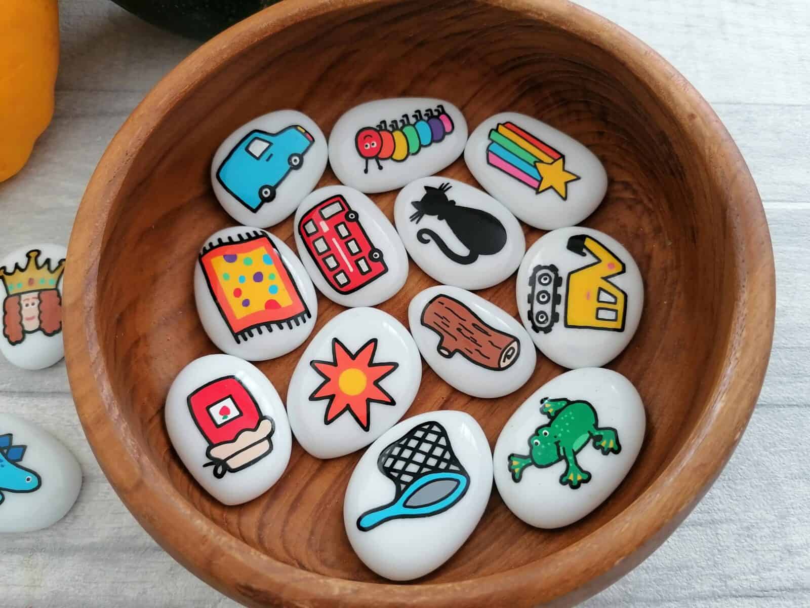 A wooden small bowl is filled with glass white pebbles with various designs painted on such as bus, sun, cat, digger, net, frog, van, caterpillar, shooting star, mat, jam and log.