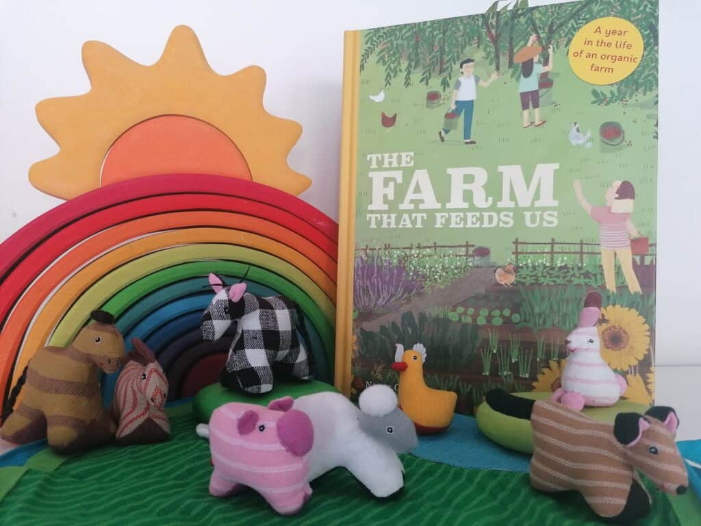 The Farm that Feeds Us book on the right with a wooden Grimms rainbow on on the left with a sun on top. Small fabric farm animals are infront.