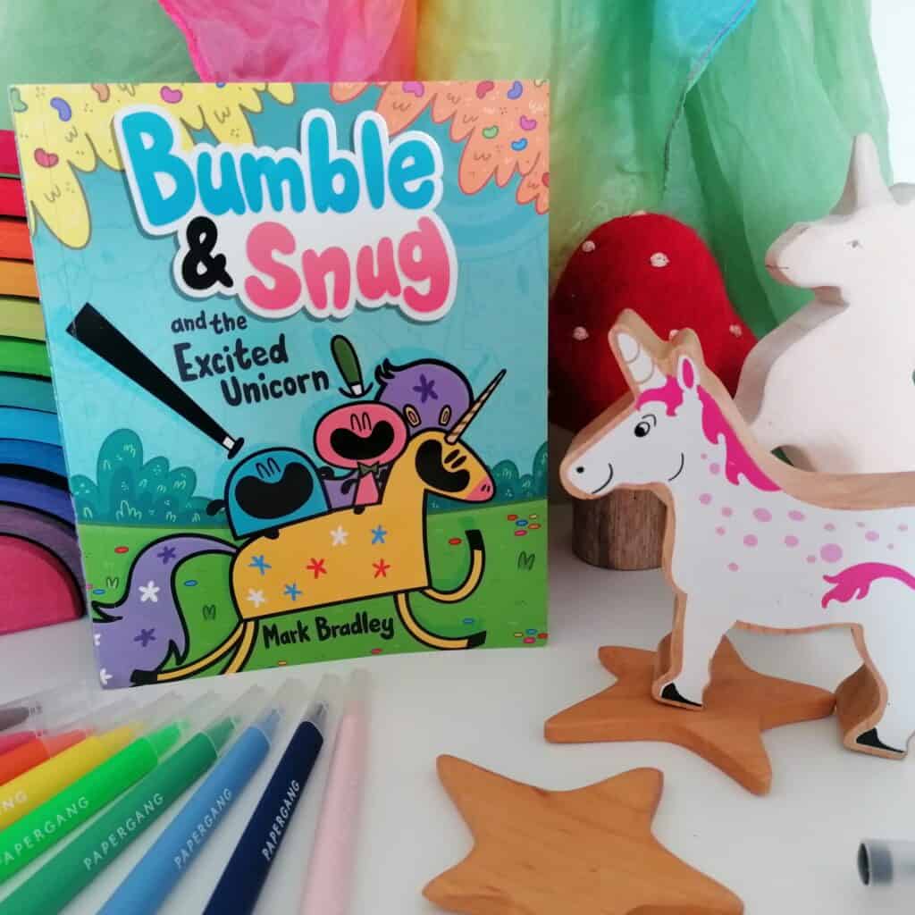 Bumble and Snug graphic novel book the Excited Unicorn front cover infront of a rainbow play silk and next to wooden unicorns.