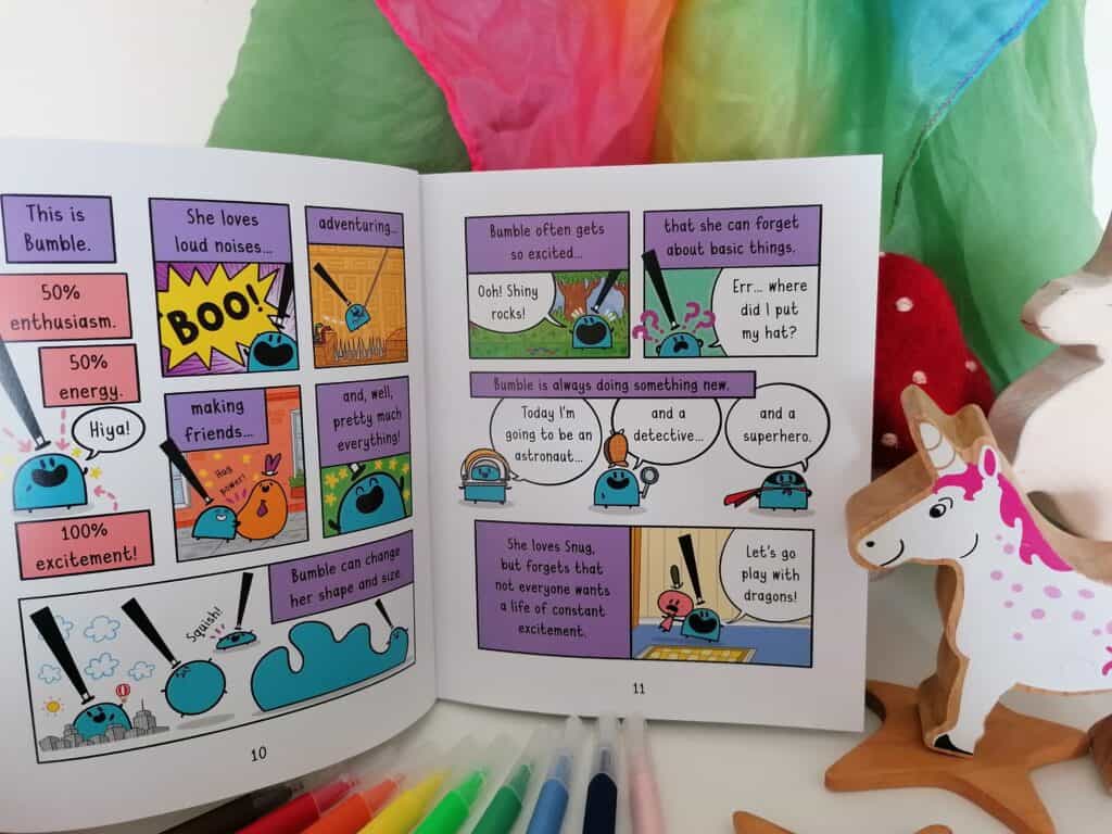 Bumble and Snug graphic novel book the Excited Unicorn inside page spread infront of a rainbow play silk and next to wooden unicorns. 